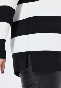 Zwart-wit gestreepte trui met een gebreide textuur, lange mouwen, zijsplitten en een rechte zoom. Draagt over zwarte broeken.