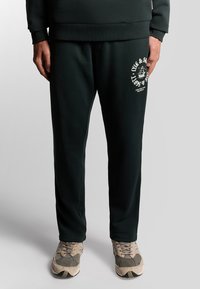 Donkergroene joggingbroek gemaakt van zachte stof met een ontspannen pasvorm, met een wit logo gedrukt op de linker bovenbeen. Gecombineerd met casual sneakers.
