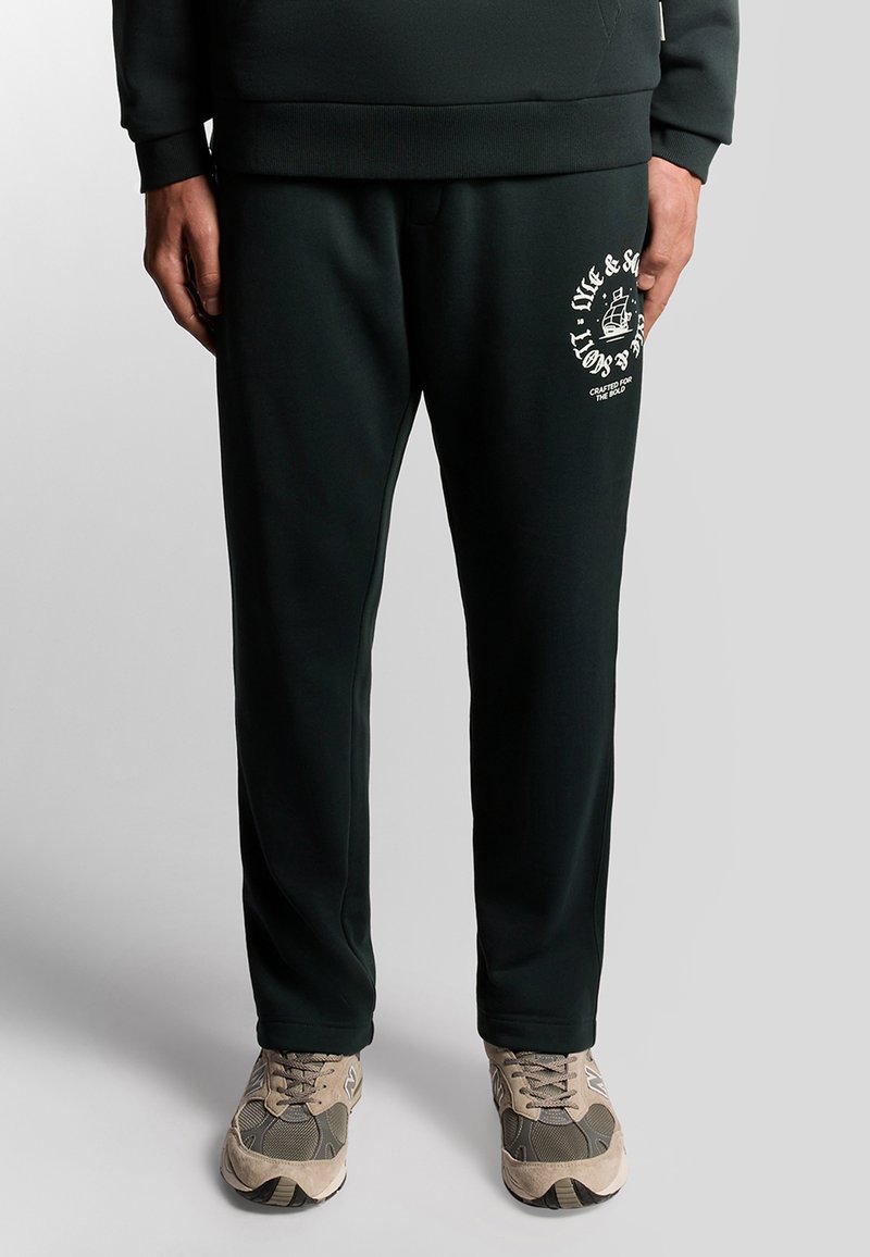 Donkergroene joggingbroek gemaakt van zachte stof met een ontspannen pasvorm, met een wit logo gedrukt op de linker bovenbeen. Gecombineerd met casual sneakers.