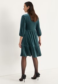 Teal fluwelen jurk met driekwart pofmouwen, tiered ruches aan de onderkant en een ontspannen fit. G gedragen met zwarte hoge hakken.