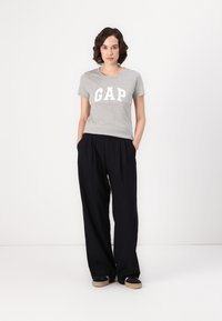 T-shirt en coton gris avec le logo « GAP » en blanc, associé à un pantalon noir ample, porté avec des baskets noires sur un fond uni.