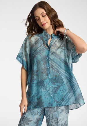 Frau mit welligem braunem Haar, die einen lockeren, transparenten blau gemusterten Knopfhemd und passende Hosen trägt, und mit der rechten Hand den Hals berührt.