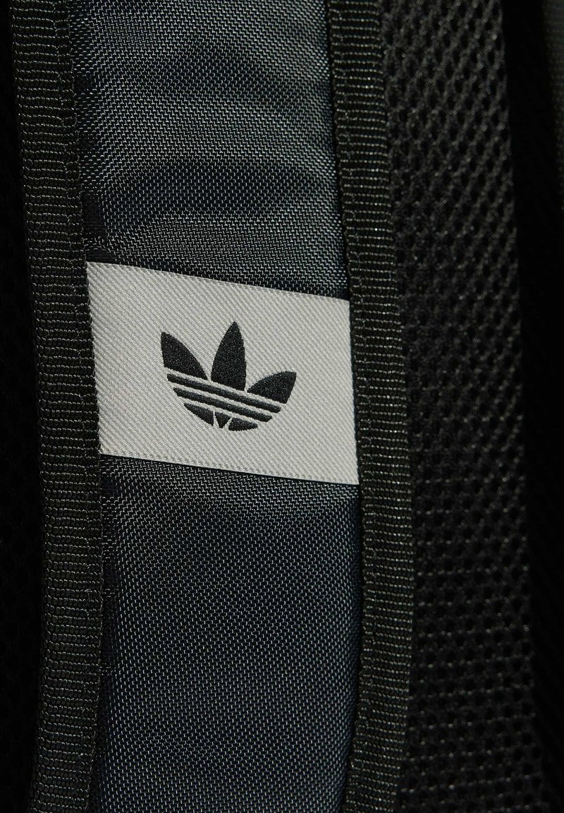 Schwarz-weißes Stoffetikett mit dem Adidas-Trefoil-Logo auf eine strukturierte dunkle Stoffoberfläche aufgenäht.