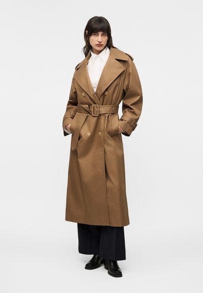 Γυναίκα που φοράει μακρύ καφέ παλτό με ζώνη πάνω από λευκό πουκάμισο, μαύρο παντελόνι με φαρδιά μπατζάκια και μαύρα μπότακια αστραγάλου, στέκεται με τα χέρια στις τσέπες.