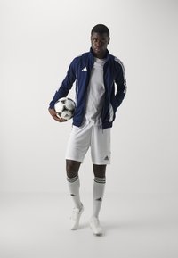 Adidas treningsantrekk med en marineblå jakke med glidelås og hvite striper, hvite shorts og fotballsko, holder en svart og hvit fotball.