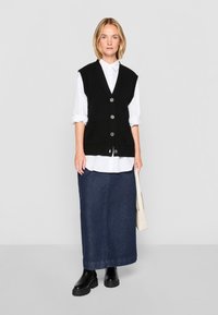 Svart ärmlös väst med knappar, vit skjorta med krage under, lång denimkjol och svarta platåstövlar; neutral bakgrund.