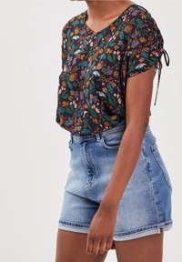 Mujer vestida con una blusa corta de manga corta de flores oscuras con lazos fruncidos, combinada con pantalones cortos de mezclilla de tiro alto en azul claro.