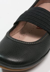 Zapato de cuero negro con punta redondeada, cinturón elástico negro y plantilla de color marrón claro con pequeños puntos. Costuras detalladas alrededor del borde.
