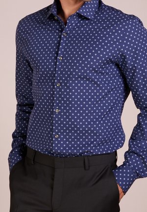 Mężczyzna w niebieskiej koszuli zapinanej na guziki z wzorem, włożonej do czarnych spodni, z rękami w kieszeniach, zwrócony lekko w lewo.