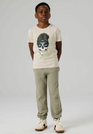 T-shirt beige avec un graphisme d'un crâne portant des lunettes de soleil et un bonnet, associé à un pantalon de survêtement vert clair et des baskets blanches avec des accents.