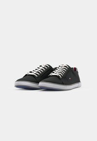 Zapatillas de lona negras con suelas de goma grises, cordones blancos y un sutil detalle de logo. Presentan un diseño de caña baja y un estilo minimalista.