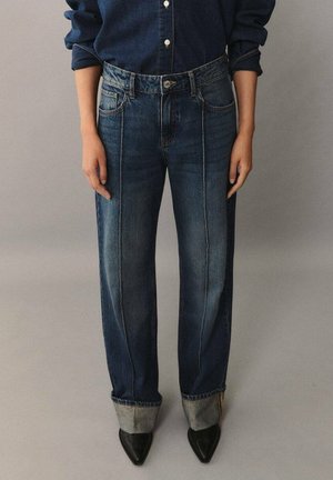 Person trägt dunkelblaue Jeans mit umgekrempelten Säumen und schwarzen spitzen Schuhen, kombiniert mit einem dunkelblauen Hemd mit Knöpfen.