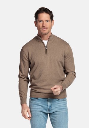 Man draagt een bruine trui met een kwart-ritssluiting over een wit overhemd en lichtblauwe spijkerbroek, staand tegen een effen witte achtergrond.