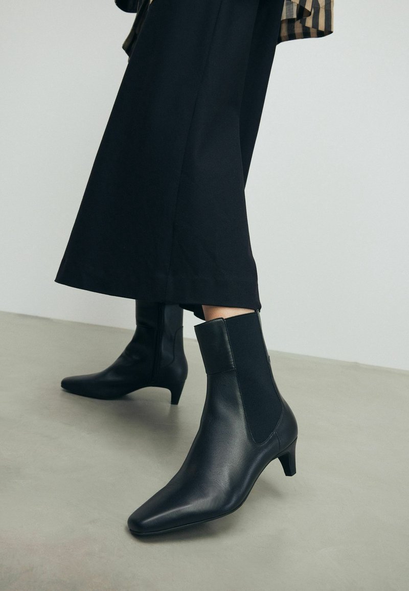 Bottines en cuir noir à talons bas portées avec une jupe noire mi-longue, debout sur un sol gris clair devant un mur uni.