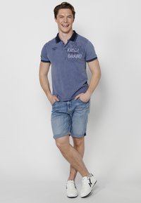 Moška modra polo majica s kratkimi rokavi, mornarsko modrim ovratnikom, grafičnim tiskom, v kombinaciji s svetlim denim kratkimi hlačami in belimi supergami. Gladka tekstura.