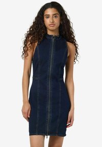 Temno denim brezrokavnik z visokim ovratnikom, sprednjim zadrgo in prilegajočo se silhueto. Ima kontrasten šiv in minimalističen dizajn.