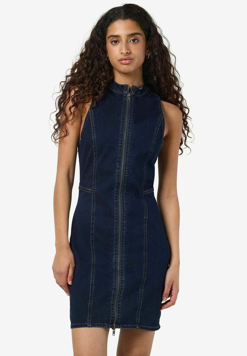 Temno denim brezrokavnik z visokim ovratnikom, sprednjim zadrgo in prilegajočo se silhueto. Ima kontrasten šiv in minimalističen dizajn.