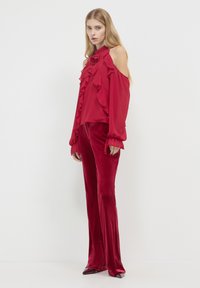 Rote Bluse mit Rüschendesign und kalten Schultern, kombiniert mit roten samtigen Schlaghosen und spitzen Absätzen. Glatte Textur und fließende Silhouette.