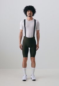Zwarte fietskorte broek met verstelbare zwarte bretels over een witte mesh top. Het model draagt witte crew-sokken en fietsschoenen.