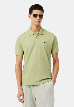 Homme portant un polo à manches courtes vert clair avec un logo de crocodile, un pantalon gris et des lunettes de soleil foncées, debout avec une main dans la poche.
