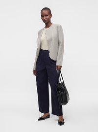 Femme portant une veste légère texturée, un top crème, un pantalon large foncé, des ballerines noires, et tenant un sac cabas en cuir noir.
