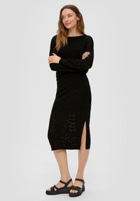 Robe noire en maille avec un motif en chevron, dotée de manches longues et d'une fente sur le côté. Associée à des sandales noires à lanières.