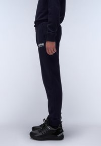 Marineblauwe sweatpants met zijzakken, een elastische tailleband en benadrukte boorden, gecombineerd met zwart sportief schoeisel met textuurelementen.