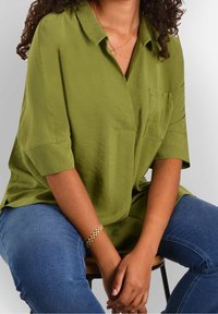 Olive green shirt with a collar, short sleeves, and a chest pocket; paired with blue jeans and a gold chain bracelet. Smooth texture.