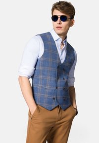 Niebieska kamizelka w kratkę z brązowymi akcentami, zapinana na podwójne guziki, noszona na jasnoniebieskiej koszuli. Brązowe spodnie i okulary przeciwsłoneczne dopełniają całość.