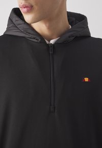 Felpa nera con cappuccio caratterizzata da una zip frontale a metà, con una superficie di tessuto liscia e un piccolo logo ricamato in rosso, giallo e nero.