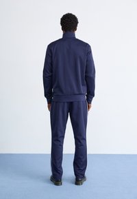 Marineblauw trainingspak met een hoge kraag, ribbels aan de mouwen en zijstrepen op de broek. Gemaakt van een soepele stof, met een loose fit en een elastische tailleband.