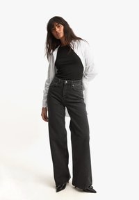 Los pantalones vaqueros de mezclilla negra de cintura alta presentan un corte relajado, un diseño clásico de cinco bolsillos y costuras en contraste. Combinados con una blusa negra y una camisa blanca.