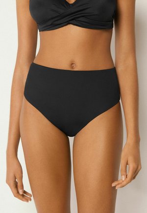 Femme portant un bas de bikini noir taille haute et un haut de bikini assorti, debout devant un fond clair uni.