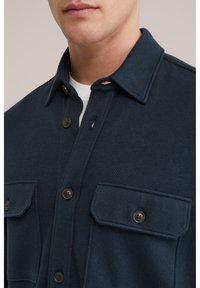 Muž nosiaci tmavomodrú textúrovanú košeľu na gombíky cez biele tričko s okrúhlym výstrihom, viditeľný golier, gombíky a náprsné vrecká.