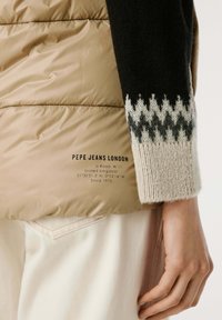Beige gewatteerd vest met zwarte gebreide mouwen, versierd met een crème- en grijze zigzagpatroon. Tekstlogo op de onderste sectie van het vest.