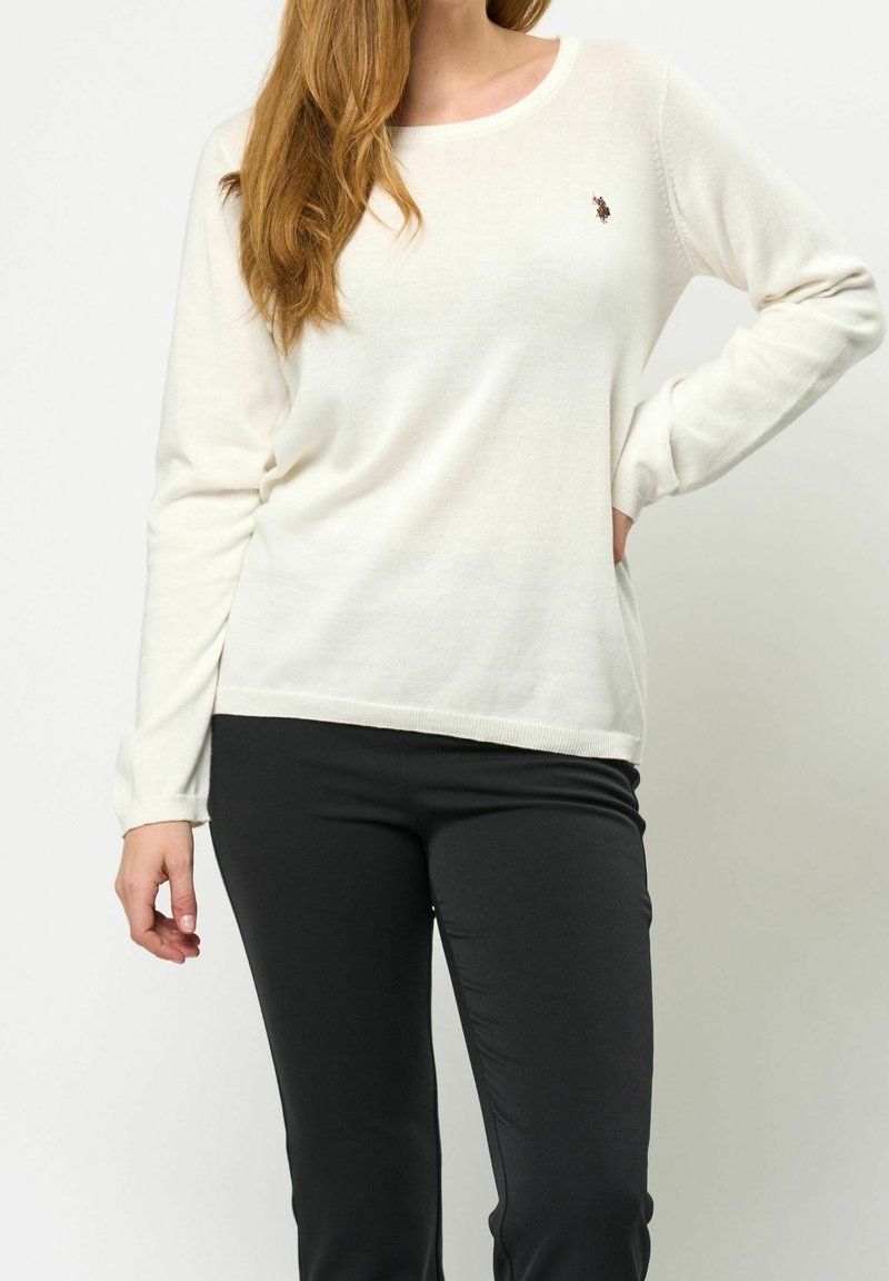 Femme portant un pull blanc à manches longues avec un petit logo sur la poitrine et un pantalon noir ajusté, debout avec une main sur la hanche.