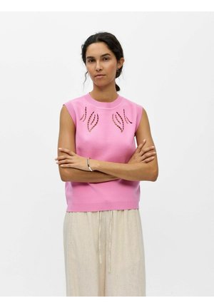 Femeie cu păr închis la culoare, purtând un top roz fără mâneci cu modele decupate în formă de frunze, pantaloni bej și o bratara argintie, stând cu brațele încrucișate.