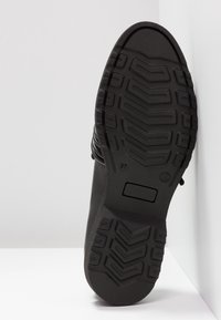 Semelle d'une chaussure noire avec des motifs de bande de roulement profonds, comprenant des formes en chevrons et des sections rectangulaires, présentée sur un fond blanc dans un coin.