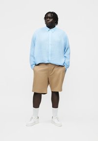 Suaugęs asmuo su tamsia oda ir trumpais dredaškais vilki šviesiai mėlyną ilgomis rankovėmis susagstomą marškinių švarką, smėlio spalvos šortus, baltas kojines ir baltus sportinius batelius, stovintis.