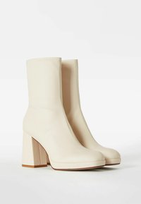 Botines de color crema con textura suave, punta redondeada y tacón grueso. Presentan costuras mínimas y un diseño sin costuras.