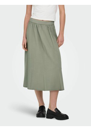 Femme portant une jupe midi taille haute en forme trapèze de couleur olive, un haut court blanc et des mocassins noirs, debout devant un fond blanc.