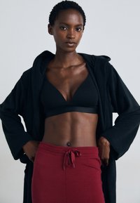 Svart hetteklær over en svart bralette og røde snorbukser. Glatte stoffer, avslappet passform, teksturert liv, og minimalistisk design.