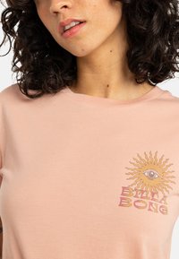Lys rosa bomull T-skjorte med rund hals, med et sol- og øye-grafikk i brun og rød på venstre brystområde.