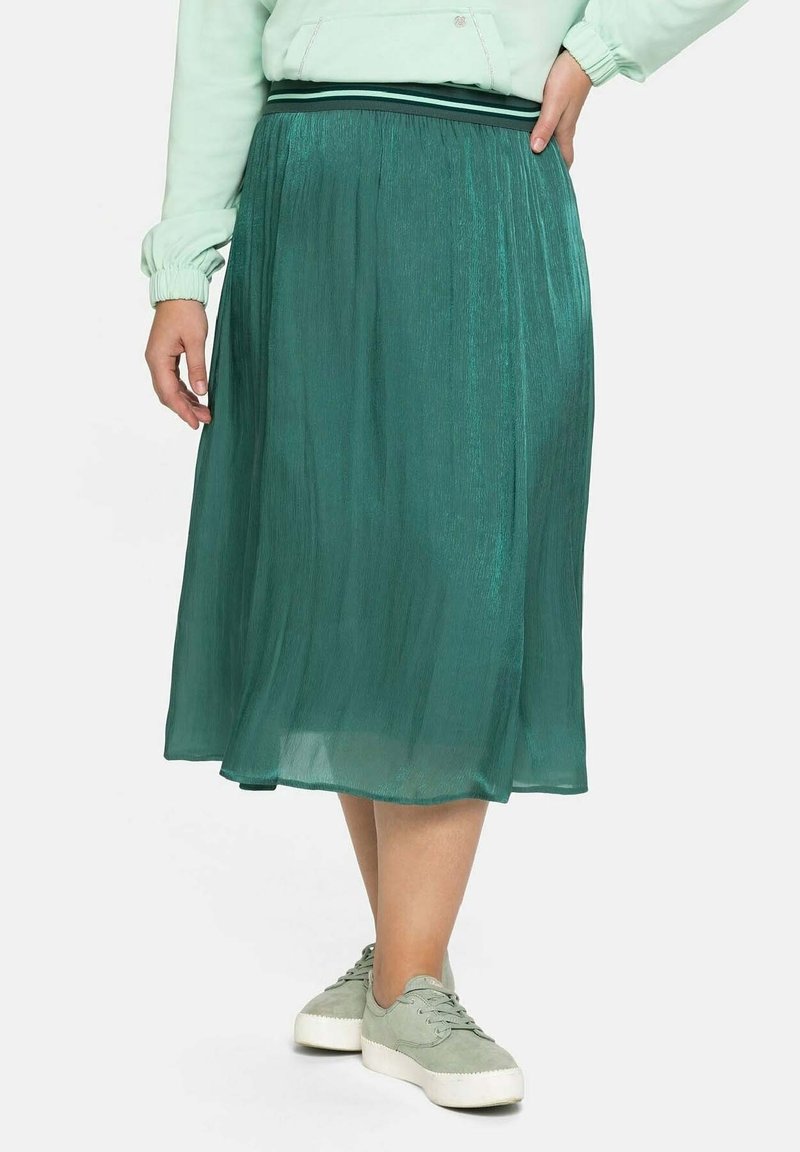 Femme portant une jupe midi plissée couleur sarcelle, un sweat-shirt vert clair et des baskets assorties, debout avec une main sur la hanche.