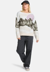 Pull en tissu tricoté avec un motif de paysage montagneux en vert, crème et rose. Associé à un pantalon sombre et des baskets.