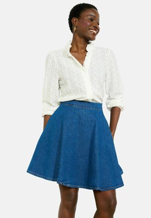 Naeratav naine, kes kannab valget silmatorkavat pluusi rollitud varrukatega ja sinist denimi lehvitavat seelikut, käed taskutes, seisab ühevärvilise tausta ees.