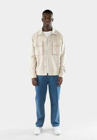 Giacca di velluto a coste beige con due tasche sul petto, t-shirt bianca sotto, jeans blu a gamba dritta e sneakers bianche.