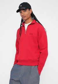 Sudadera roja con cremallera y cuello alto, con texto bordado. Combinada con pantalones grises de corte relajado y dobladillos ajustados.