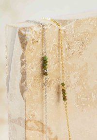 Dos collares en capas exhibidos sobre una superficie de piedra texturizada. Uno tiene una cadena de plata con cuentas verdes, el otro tiene una cadena de oro con cuentas similares.