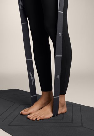 Sorte leggings, bare fødder på en mørk yogamåtte. To grå modstandsbånd med markeringer er placeret lodret ved siden af fødderne.